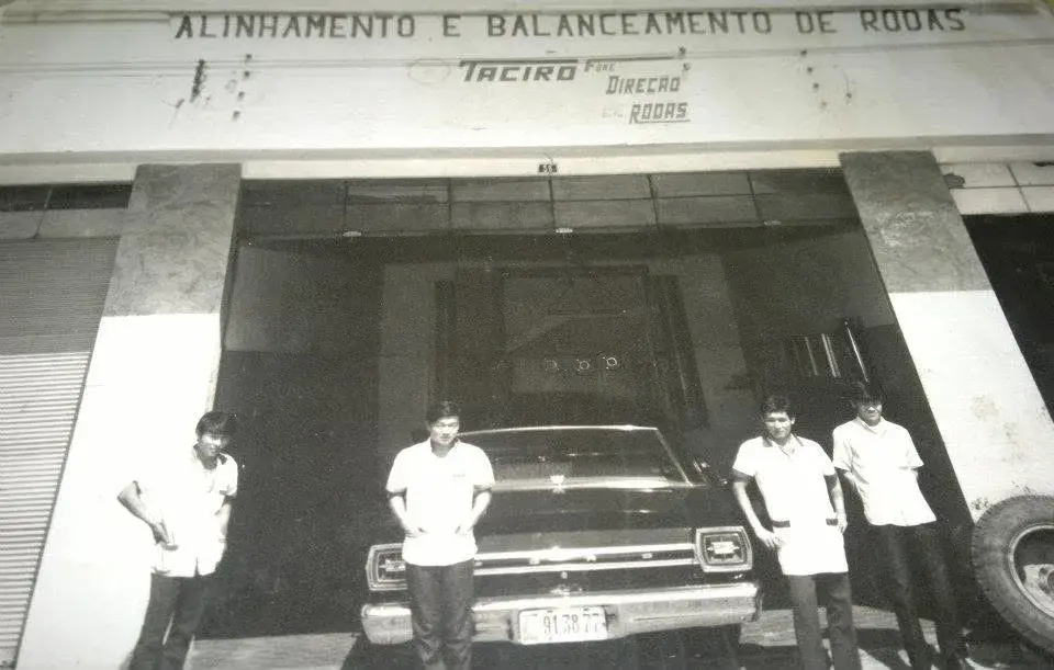 Fachada antiga da Taciro em preto e branco, com o letreiro Alinhamento e Balanceamento de Rodas, quatro rapazes de uniforme claro na calçada e um Ford preto estacionado na porta