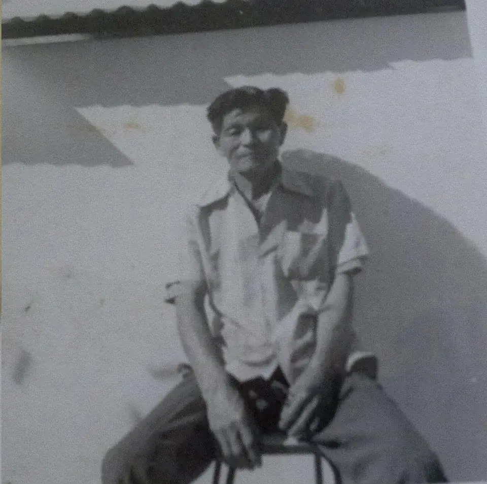 Retrato em preto e branco de um senhor de camisa clara, sentado num banquinho diante de uma parede branca