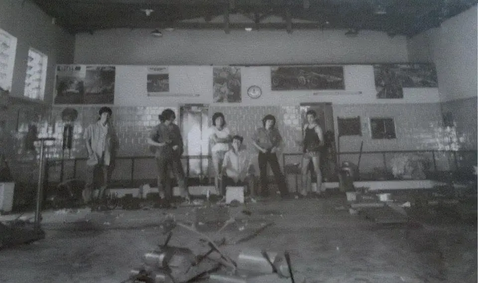 Interior antigo da oficina em preto e branco: seis rapazes, um deles sentado, diante de uma parede de azulejos com pôsteres de carros e um relógio, e o chão ainda cheio de peças