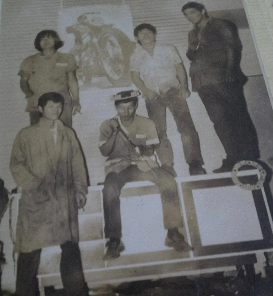 Cinco rapazes da oficina posando em cima e em volta de uma bancada, com a roupa suja de graxa e o pôster de uma moto na parede, numa foto amarelada pelo tempo