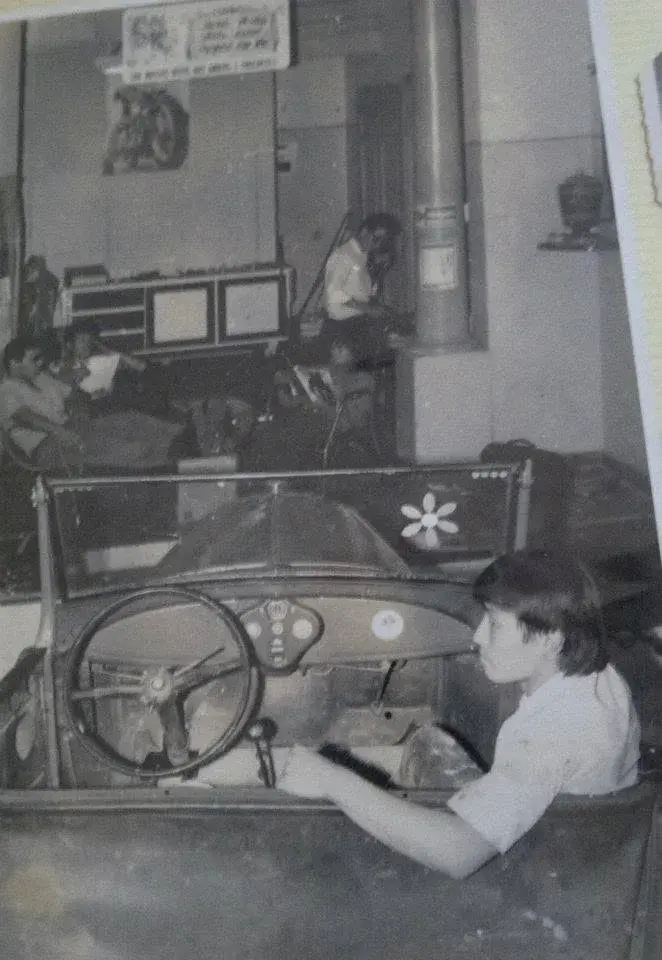 Rapaz ao lado de um carro antigo sem capota, com um adesivo de flor no para-brisa, e ao fundo a oficina com gente trabalhando e o pôster de uma moto