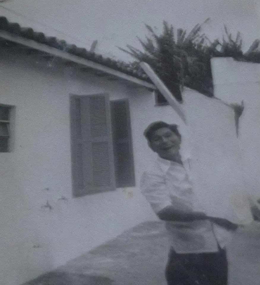 Retrato em preto e branco de um homem sorrindo, carregando uma tábua na frente de uma casa de janelas de madeira