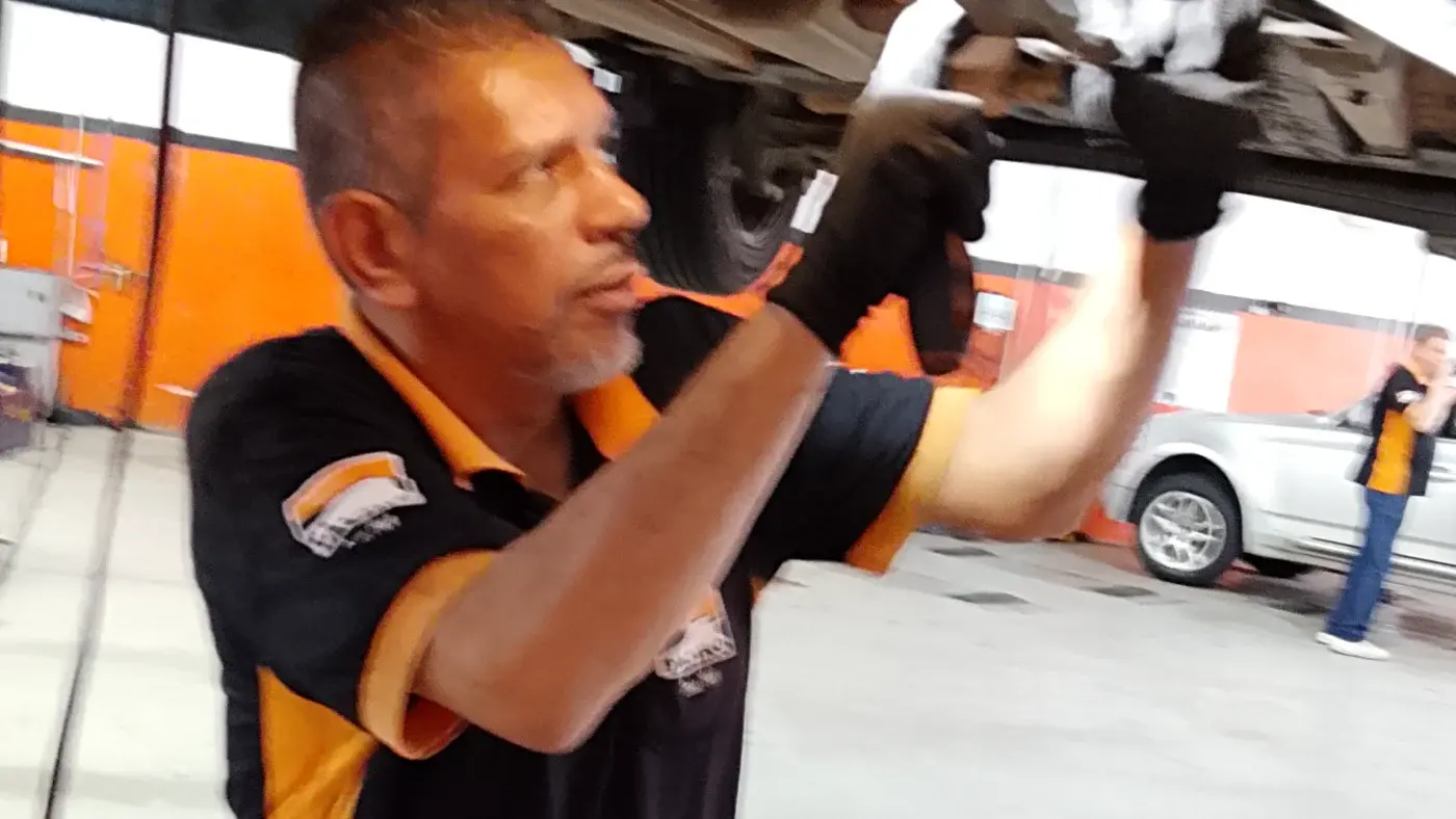 Mecânico da Taciro de perfil, com as duas mãos trabalhando embaixo de um carro suspenso, com a parede laranja da oficina ao fundo