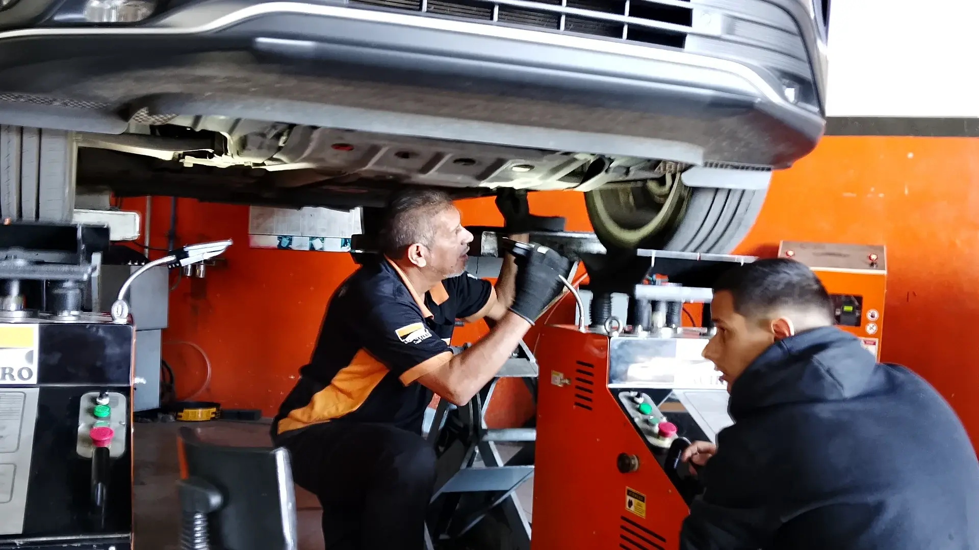 Mecânico da Taciro olhando de perto a suspensão de um carro suspenso, com a máquina de diagnóstico ao lado