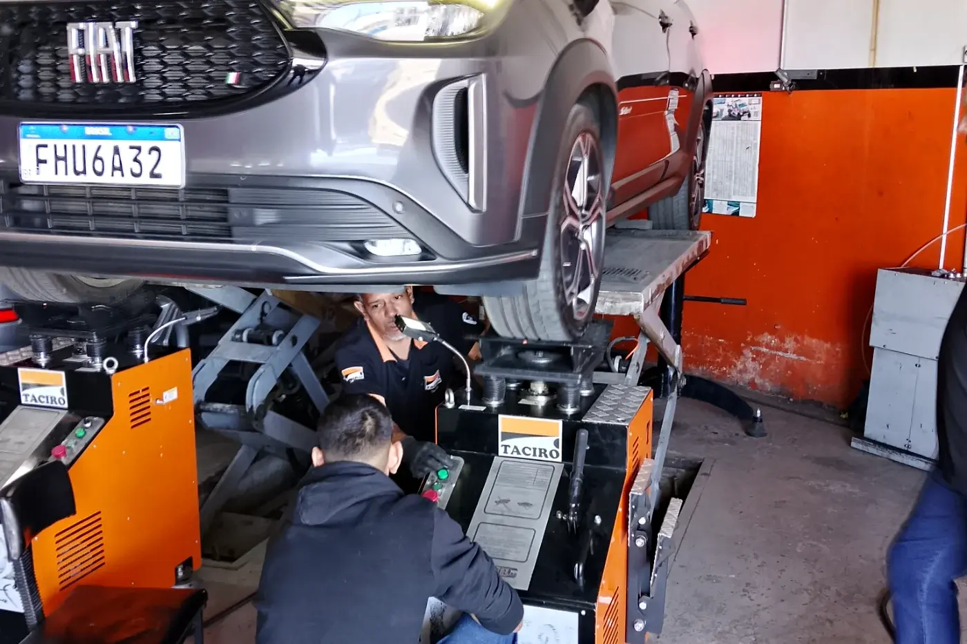 Dois mecânicos da Taciro trabalhando juntos embaixo de uma Fiat Fastback suspensa no elevador tesoura, com a máquina de balanceamento ao lado
